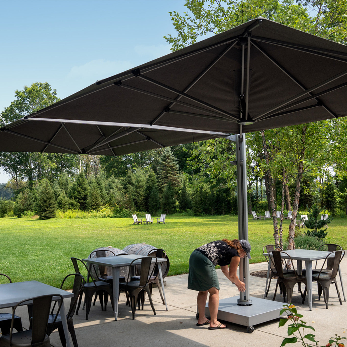 Person setting up outdoor furniture under a large black umbrella in a park-like setting.