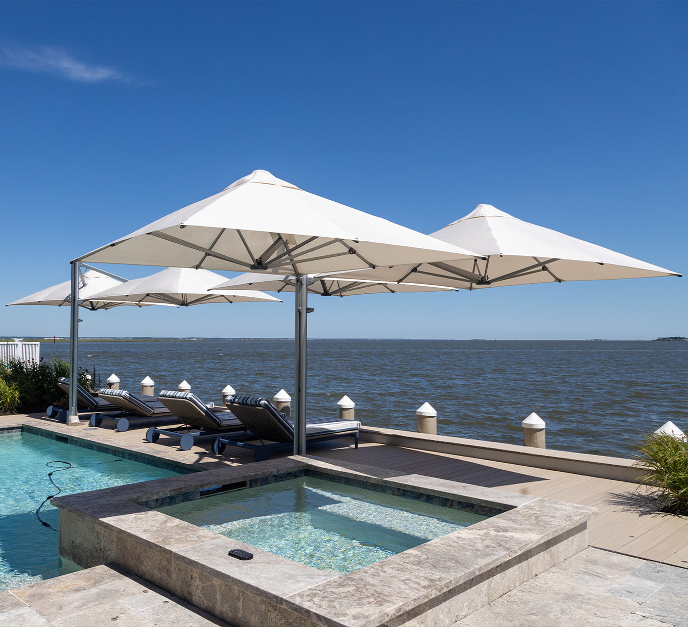 Outdoor setting with pool, lounge chairs, and large umbrellas by a body of water.