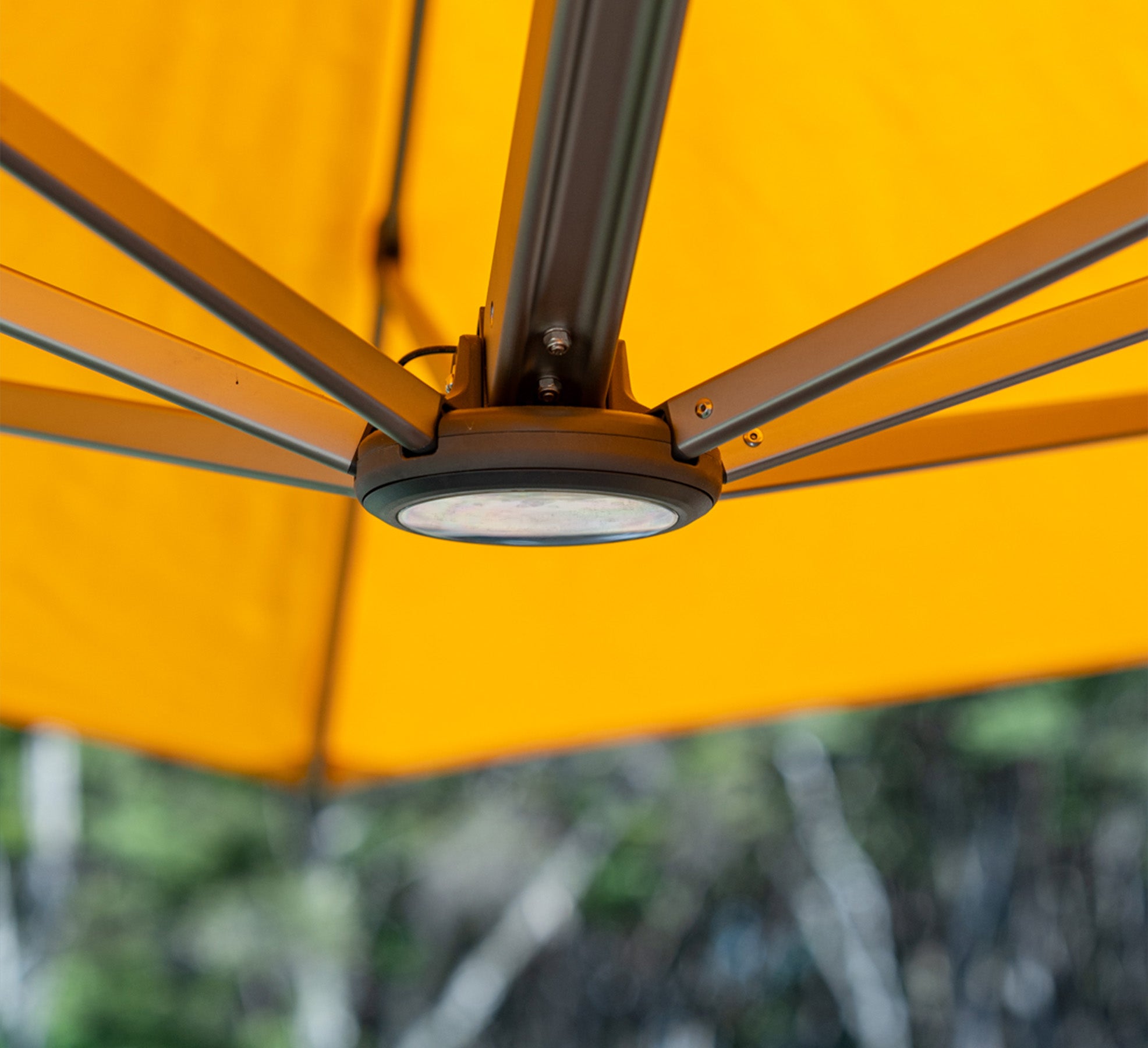 Close-up of a yellow outdoor umbrella with a light fixture, blurred greenery in the background.
