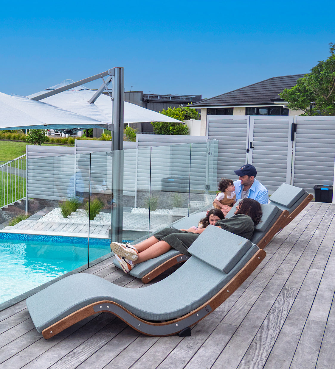 Family relaxing on modern outdoor lounge chairs by a pool with a clear blue sky.