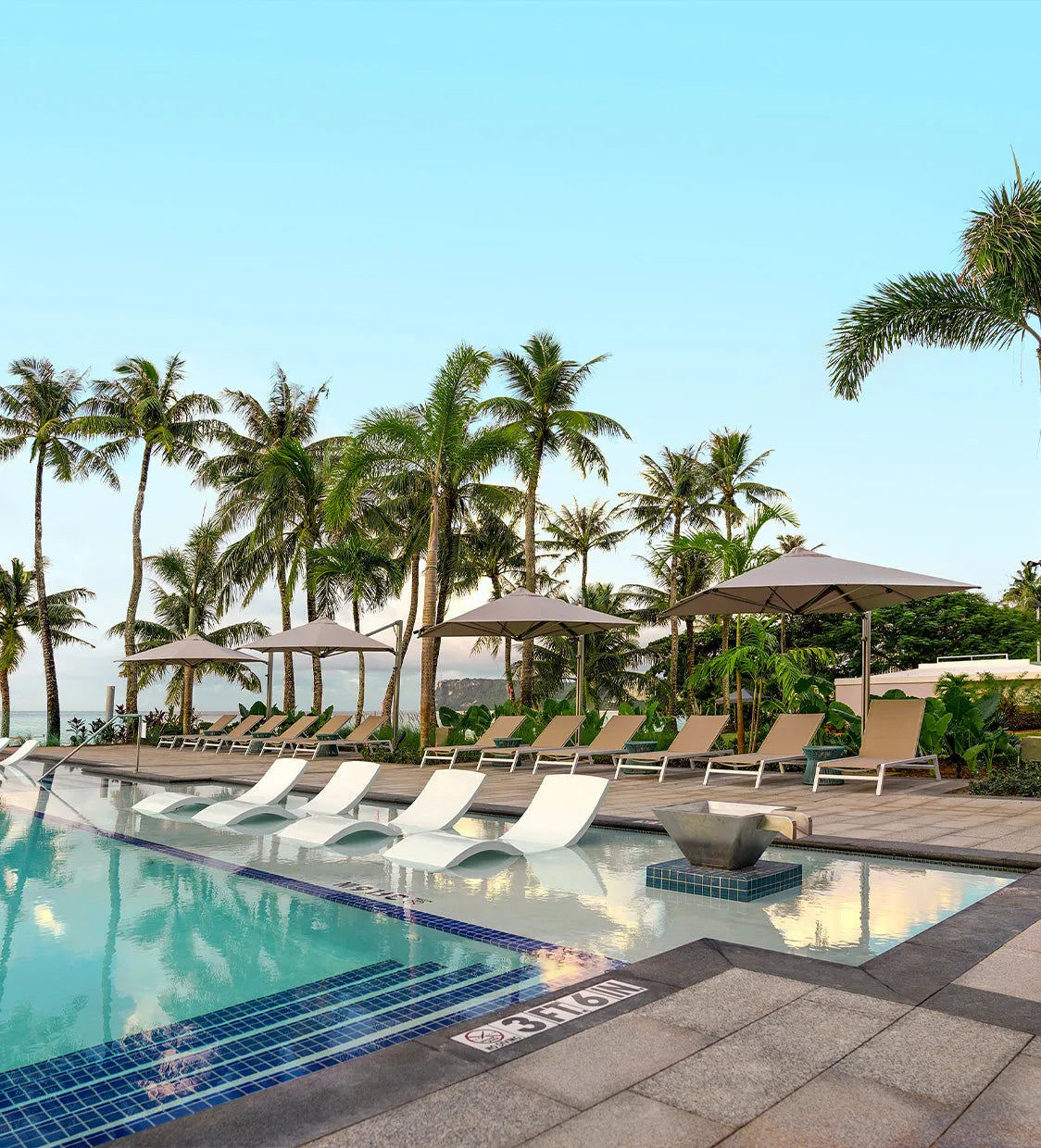 Pool area with lounge chairs, umbrellas, and palm trees under a clear blue sky.