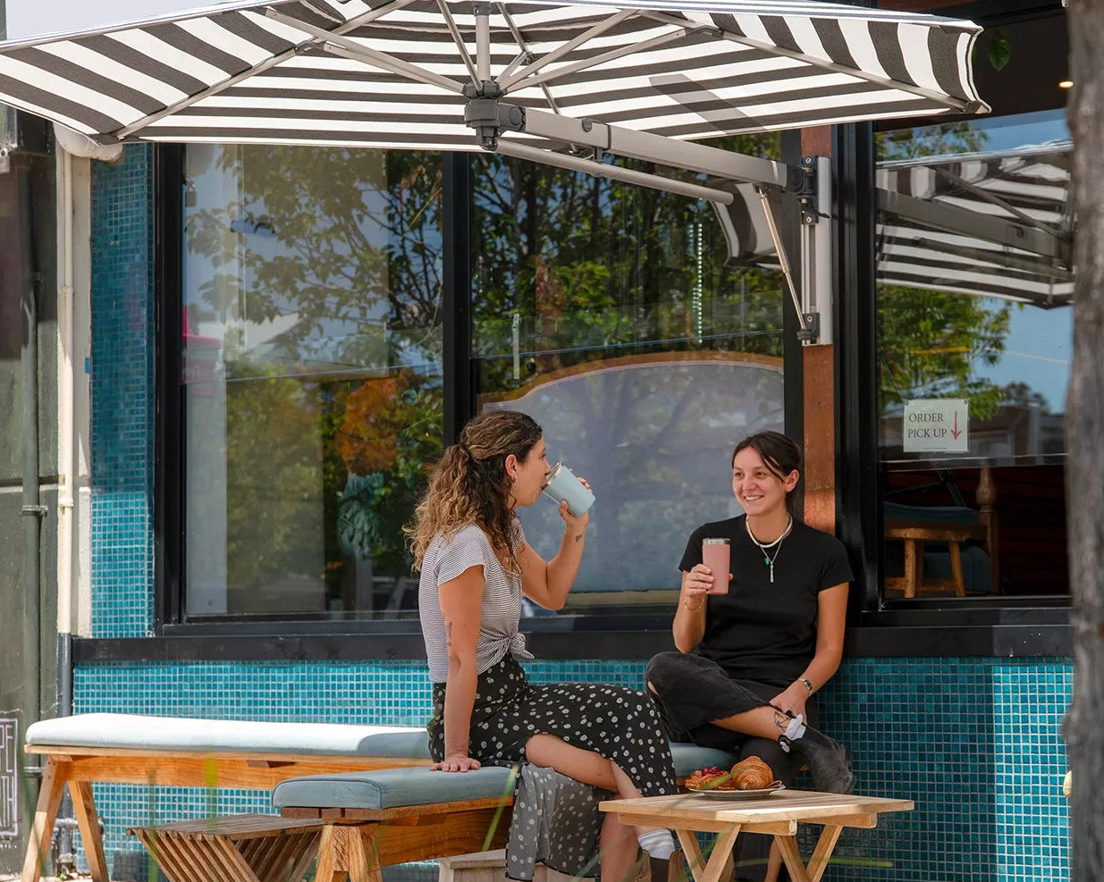 Two women sitting at a table outside a cafe with a striped umbrella and tiled wall.