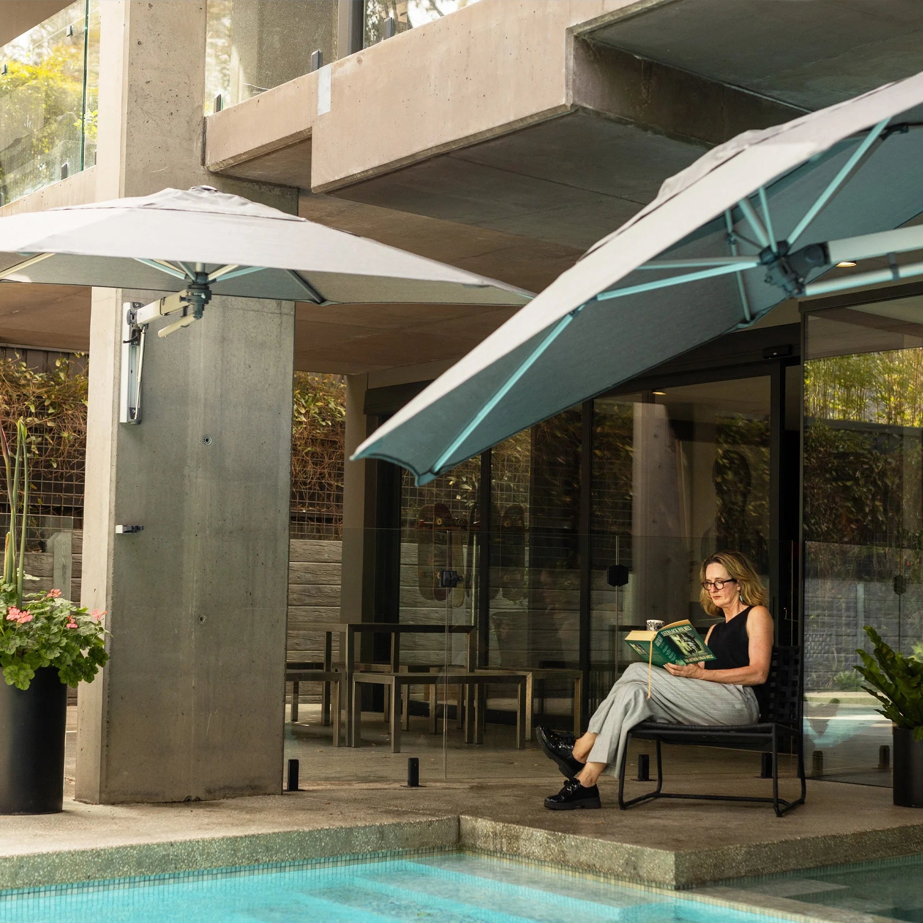 Person sitting on a chair under a large outdoor umbrella by a poolside.