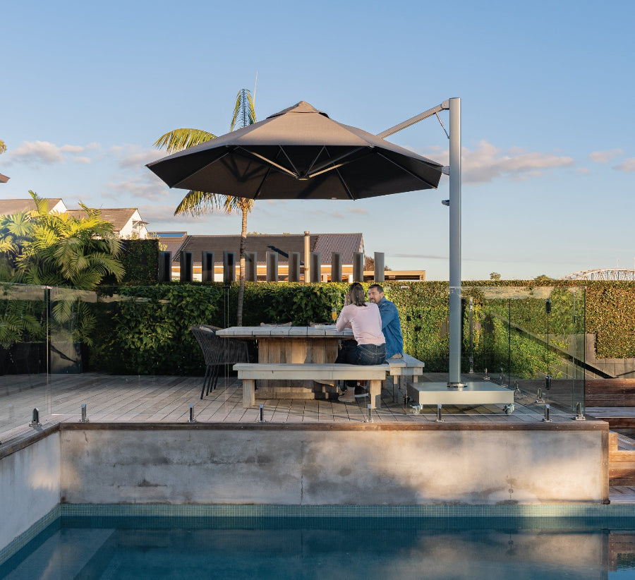 Two people sitting at a table under an umbrella by a pool with a scenic background