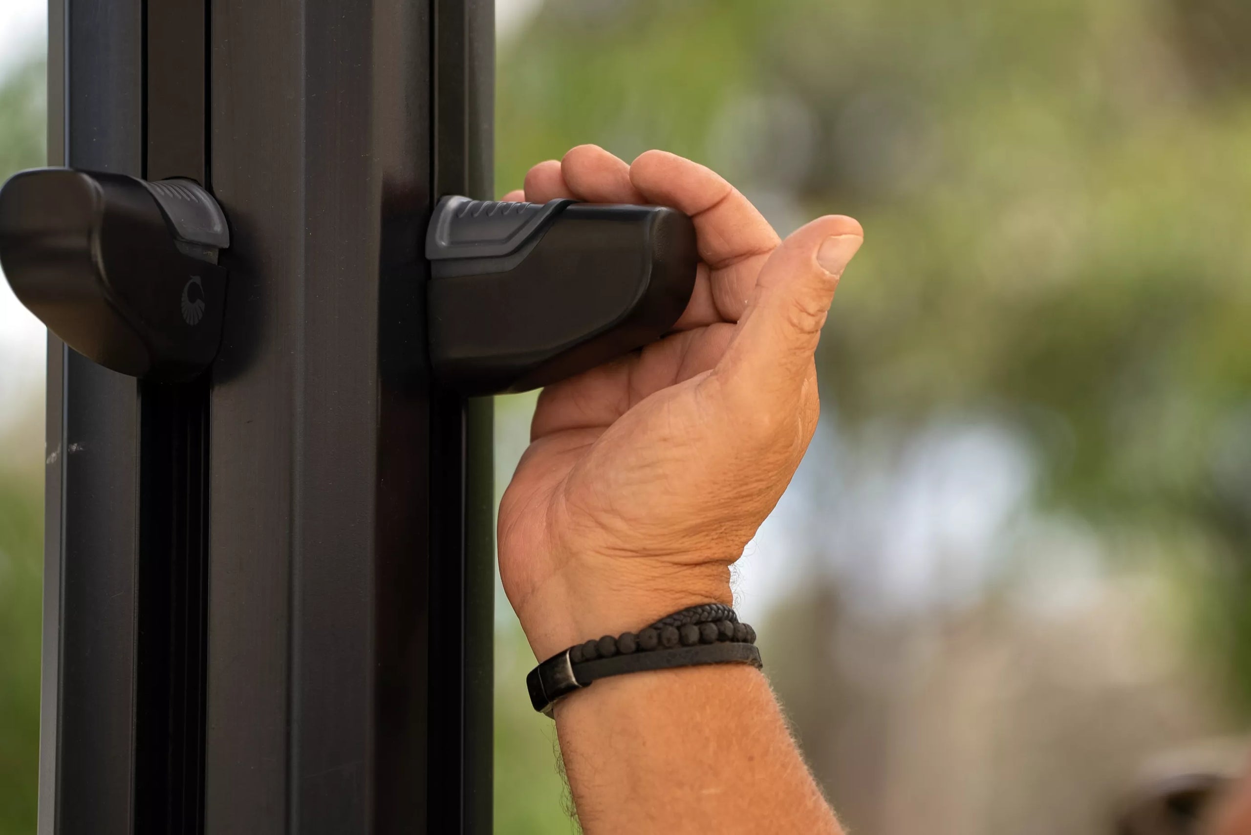 Hand opening a door with a black handle, blurred greenery in the background